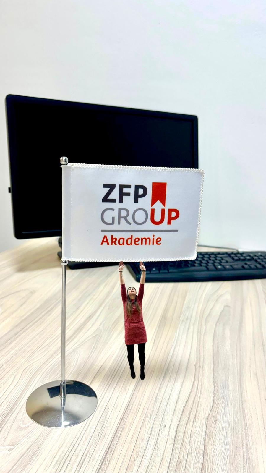 Žena v červených šatech drží transparent „ZFP GROUP Akademie“ na pódiu před notebookem.